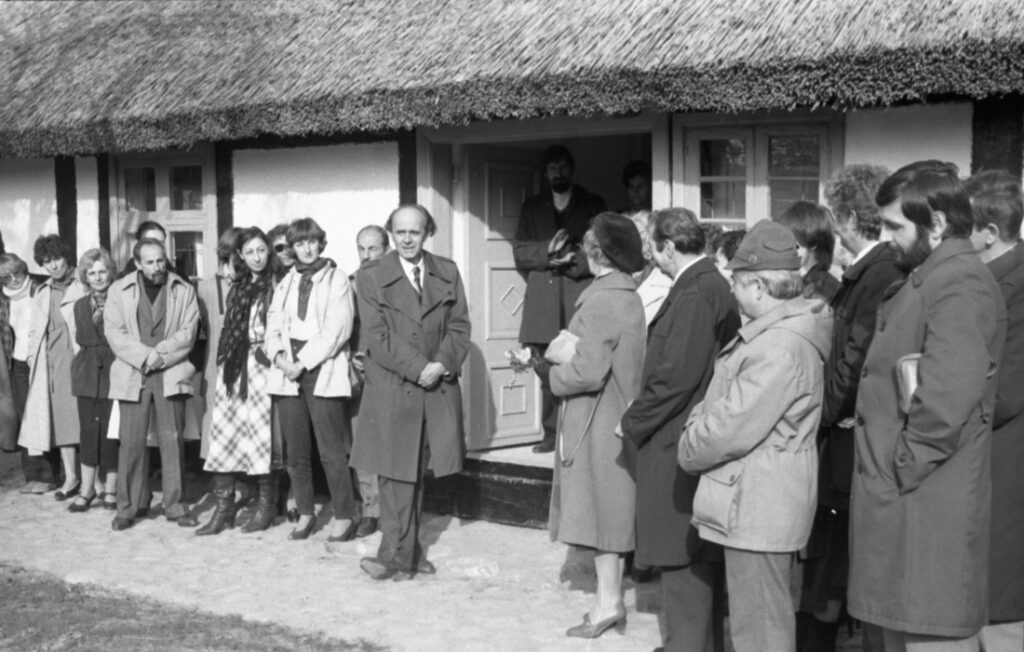 Przed chatą stoi grupa ludzi, która uczestniczy w otwarciu skansenu (kliknięcie spowoduje powiększenie zdjęcia)